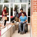 Un élève en fauteuil roulant utilise une rampe d'accès devant son établissement d'enseignement, entouré d'autres élèves qui discutent en petits groupes.
